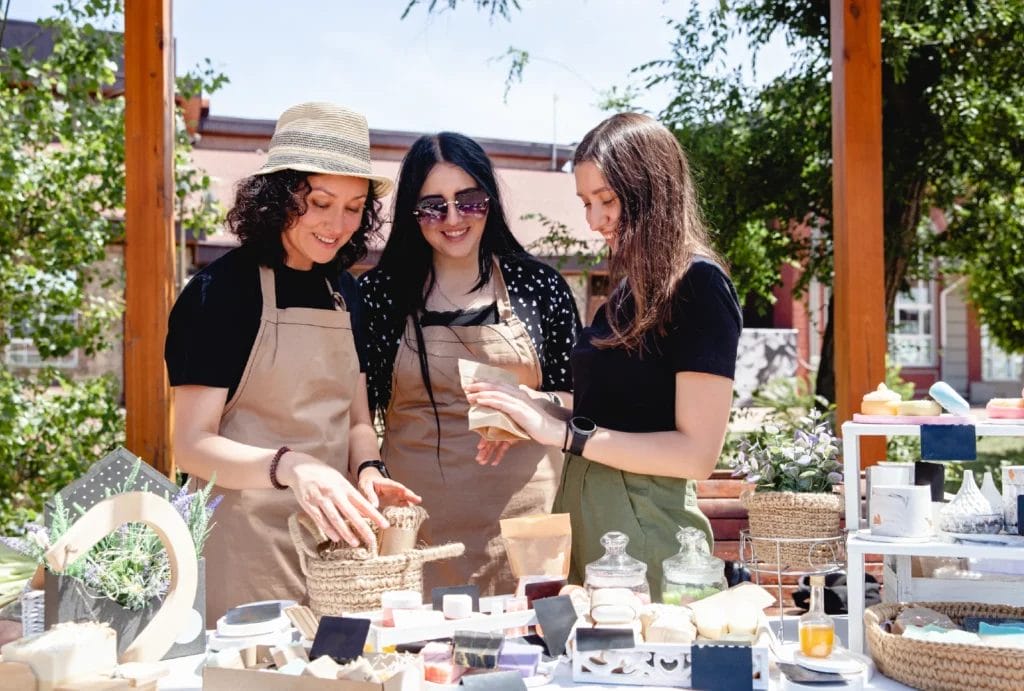 Three vendors smile and check their products at an outdoor market behind their display table with various beauty products laid out atop it.