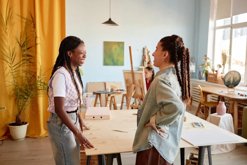 Two artists, one wearing a purple checkerboard vest and denim, and the other wearing a denim shirt and brown pants, smile and converse in a brightly lit art studio with an orange curtain in the background.