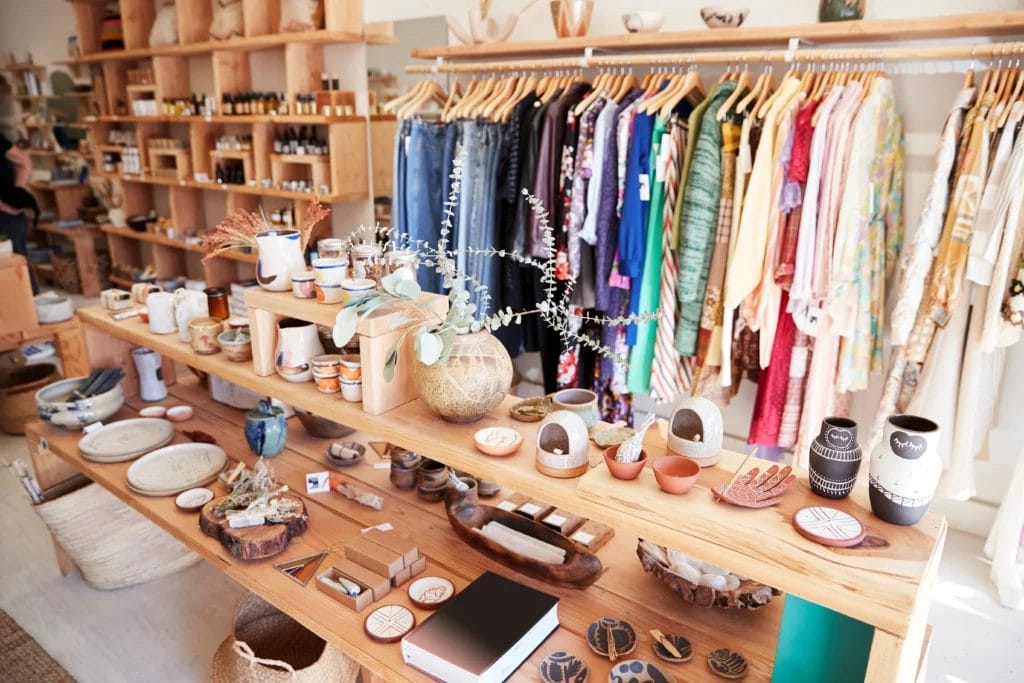 A gift store displaying craft goods on a wooden display shelf with colorful clothing hanging on a rack in the background.