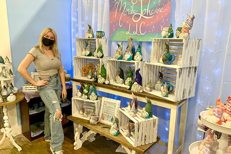 Marika stands next to a display of crates full of her colorful hand painted pottery at an event.