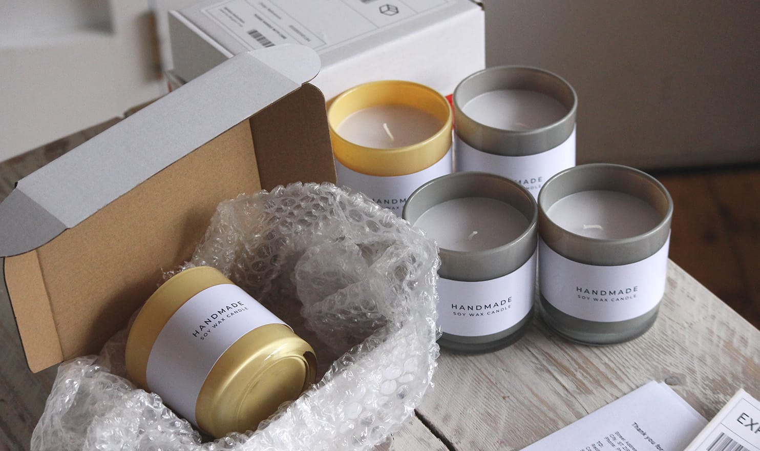 A collection of handmade soy wax candles sit on a wood table with one lying in a small shipping box with bubble wrap awaiting a label for shippment.
