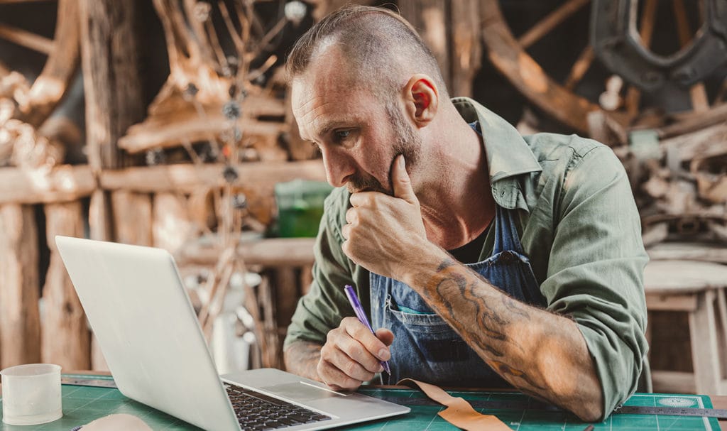 An artisan is very deep in thought as he works on his laptop in his workshop.
