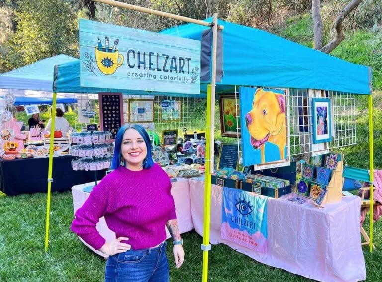Chelsea (aka Chelzart) happily standing in front of her colorful booth display at an outdoor craft fair.