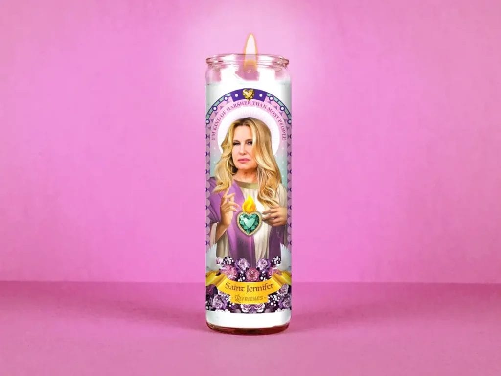 A white candle with a label that depicts Jennifer Coolidge illustrated as a saint with text that reads, "I'm kind of harsher than most people" and "Saint Jennifer, Lit Friends."
