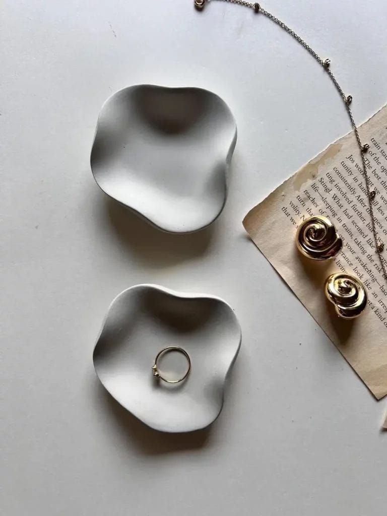 A set of white minimalist, organic-shaped jewelry trays, a worn book page, and goldtone jewelry laid out on a white table.