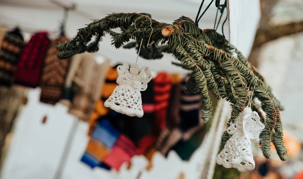 Crocheted angel Christmas ornaments hang from a fir branch while on display in a vendor booth at a craft fair.