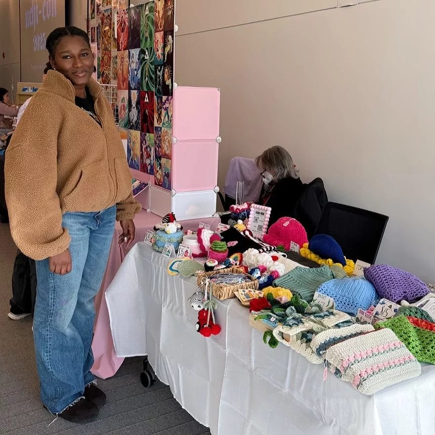 Maya, the owner of HookedUp, standing next to her crochet booth at a craft fair.