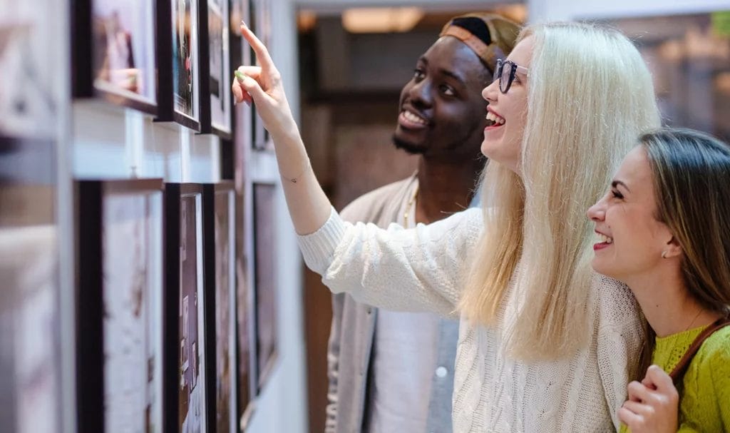 Three young people laughing while looking at a picture on display in a gallery. The girl in the middle of the group points very closely at a framed image, almost touching it.