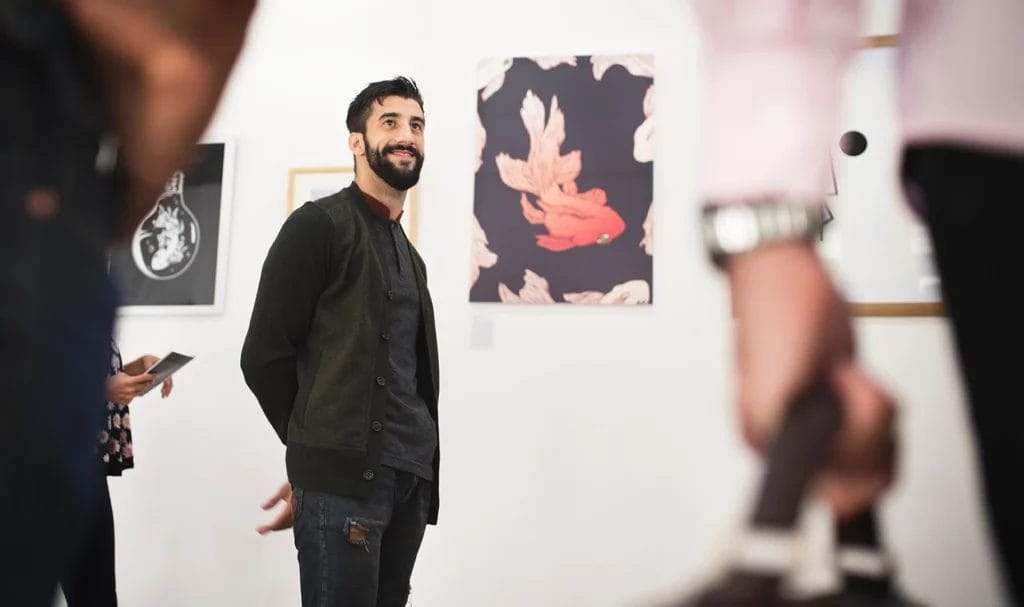 An artist proudly smiles and stands next to his work in front of a crowd of patrons attending his art exhibition.