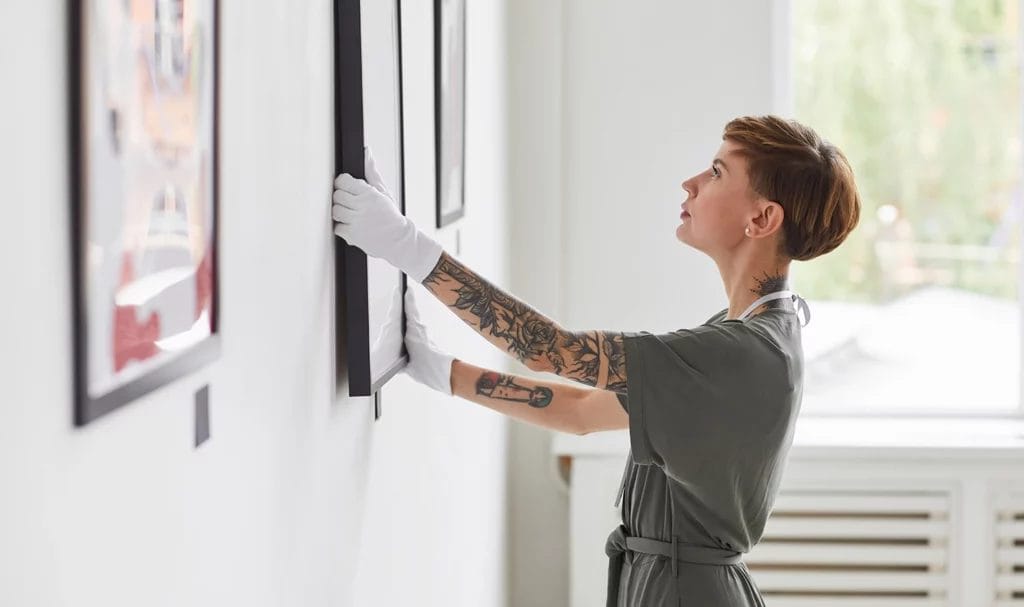 Side view portrait of tattooed creative woman hanging paintings on wall while planning art gallery exhibition.