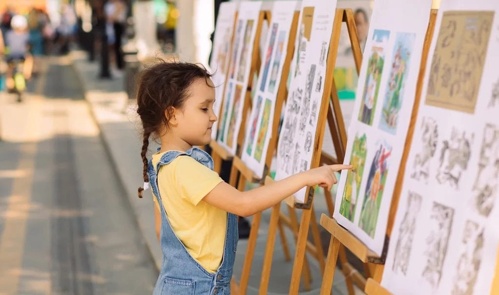 A little kid touches artwork on display on easels at an outdoor art exhibit.