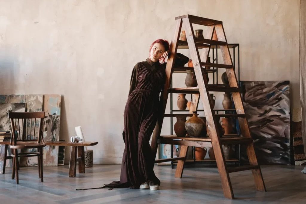 An artist wearing a brown maxi dress leans against a wooden ladder in an art studio with various paintings and earthenware jars placed throughout.