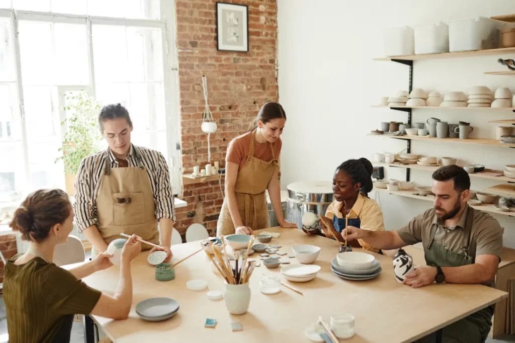 Two artists instruct a group of attendees on painting ceramics during a workshop in a ceramics studio.