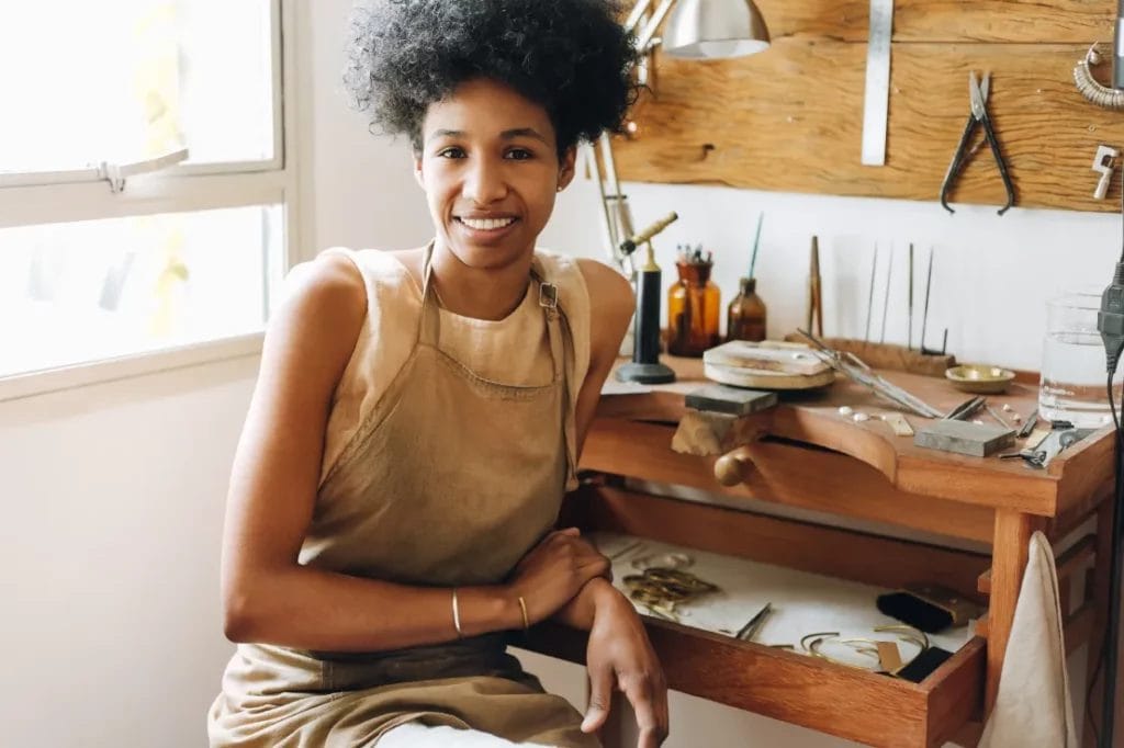 An artist wearing a beige tank top and a taupe apron smiles and leans against her wooden desk, which is covered with jewelry-making tools, in her art studio.