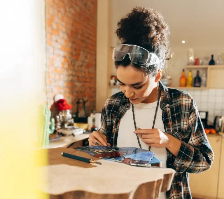An artist wearing protective eye goggles, a white t-shirt, and a navy and orange flannel works on a mosaic piece in her art studio.