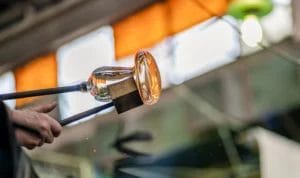 Close-up on the hand of a glassblower artist using tools to shape molten glass extracted from a furnace.