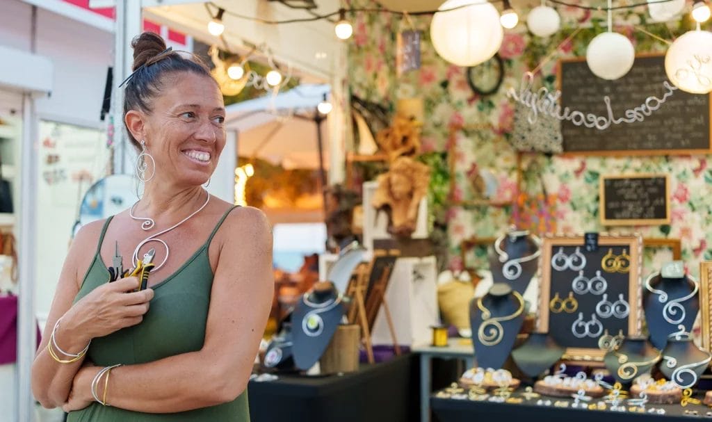 Woman artisan holding pliers smiling in front of her jewelry stall at a market.