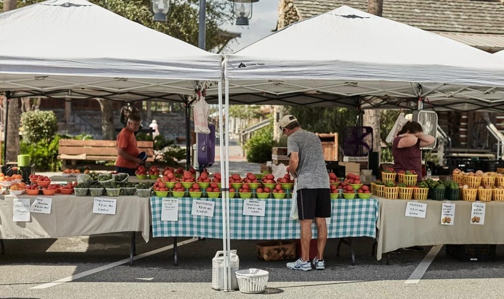 A young man purchases fresh produce from a table under some pop-up tents at an open-air street market.