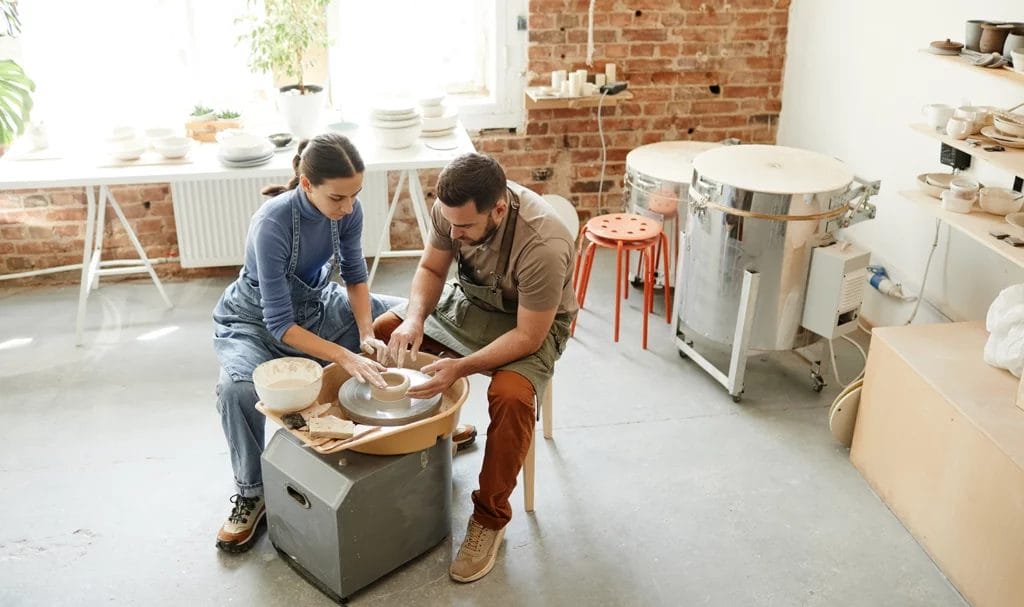 High angle photo of young couple sitting by pottery wheel together in handmade ceramics workshop.