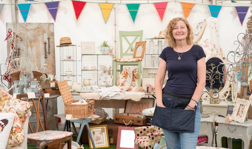 Woman smiling in front of her vintage crafts stall at a market.