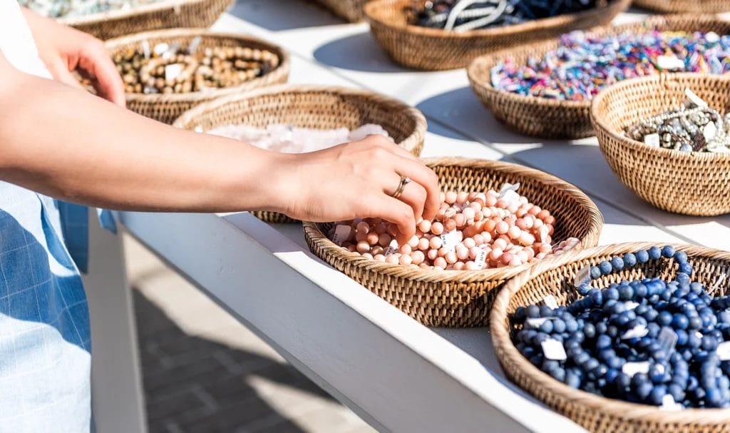 Close-up of a young woman shopping for colorful stone beach bracelets as she touches the handmade jewelry on display on a table in an outdoor market.