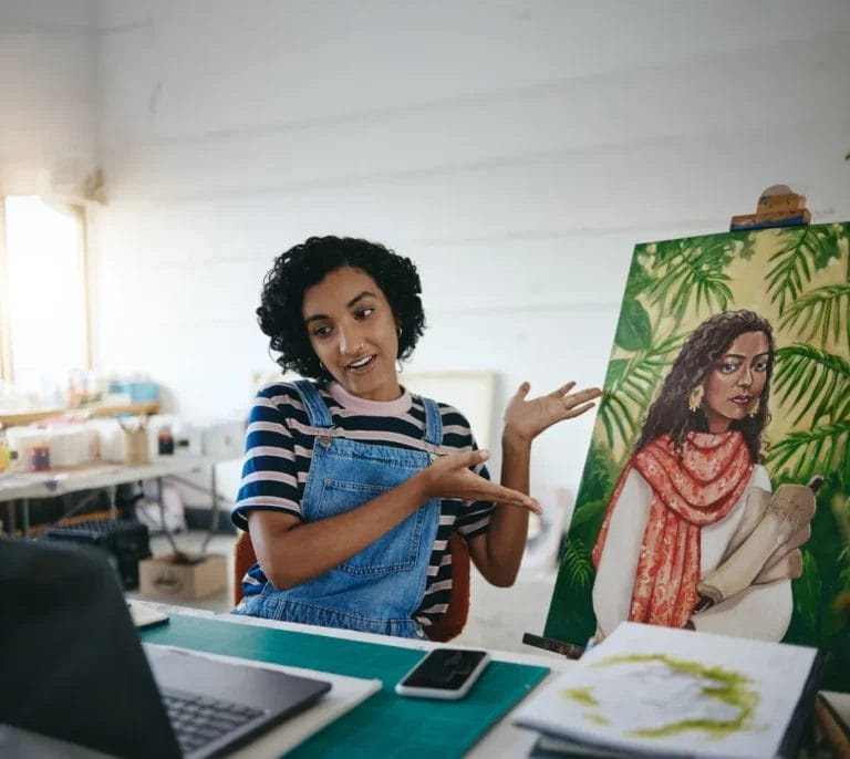A young woman with short curly hair, wearing a striped t-shirt and denim overalls, sits at a desk in front of a laptop and gestures to an art piece behind her depicting a woman surrounded by green foliage.