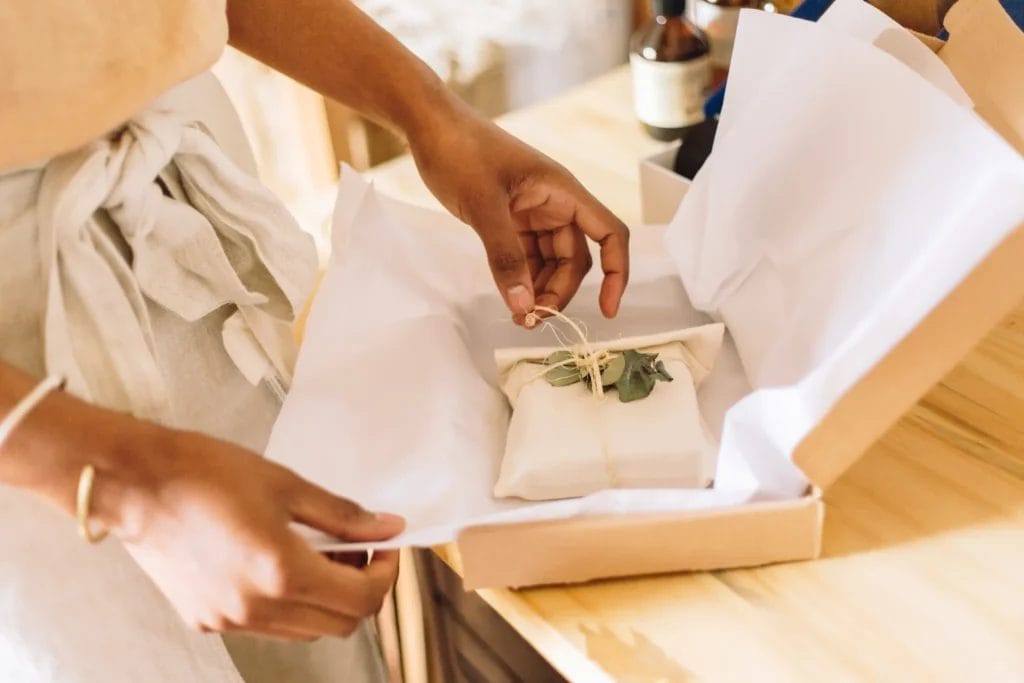 A close-up image of an artist packing an order with white tissue paper, twill, and green leaves into a cardboard box.