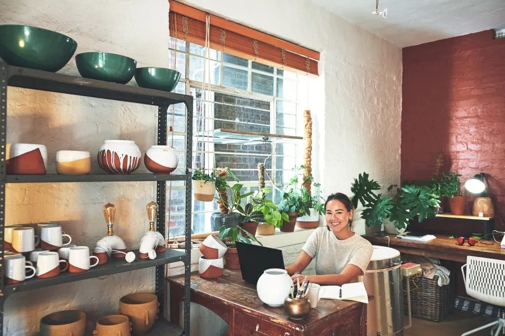 A woman smiles while typing on her laptop while sitting at a desk in a pottery studio.