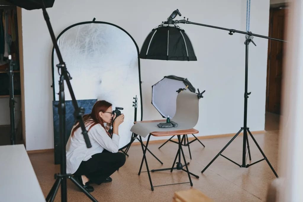 A woman crouches to take a photograph of art pieces placed on a taupe-colored backdrop draped over a wooden stool. In the studio are various lighting fixtures and reflectors.