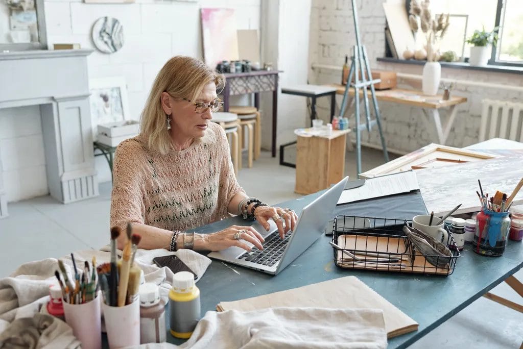 A female artist types at her laptop at a desk in her art studio. On the desk are cups of paintbrushes, canvas, and wooden frames. In the background, there are stools, an easel, potted plants, and a glass window.