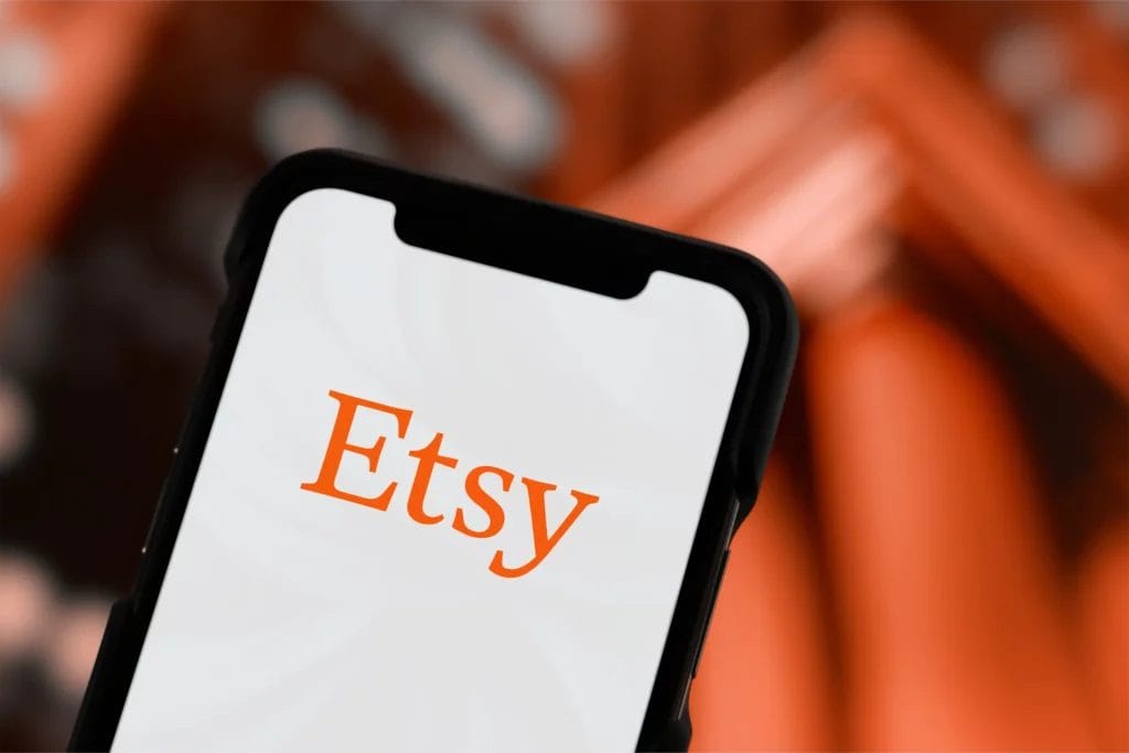 A smartphone displaying the Etsy logo above a blurred orange background.
