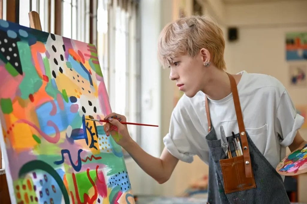 An artist wearing a white shirt and a brown-and-blue apron paints a brightly colored abstract piece on canvas in an art studio.