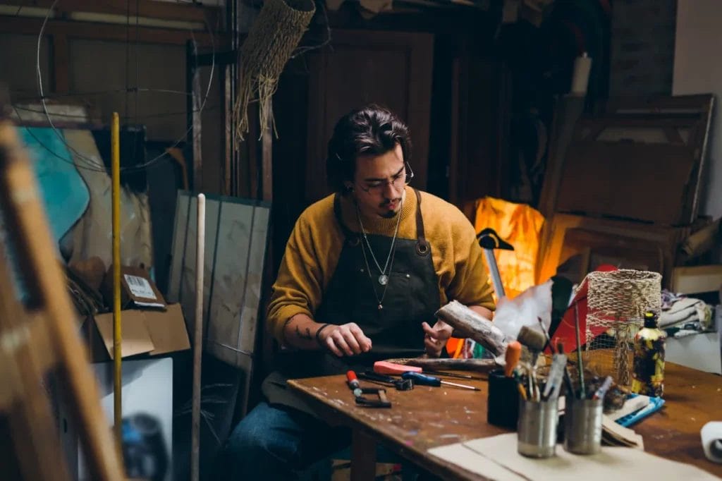 An artist wearing a mustard-colored sweater and a black apron works on a wood piece at a wooden table in a dimly lit studio.