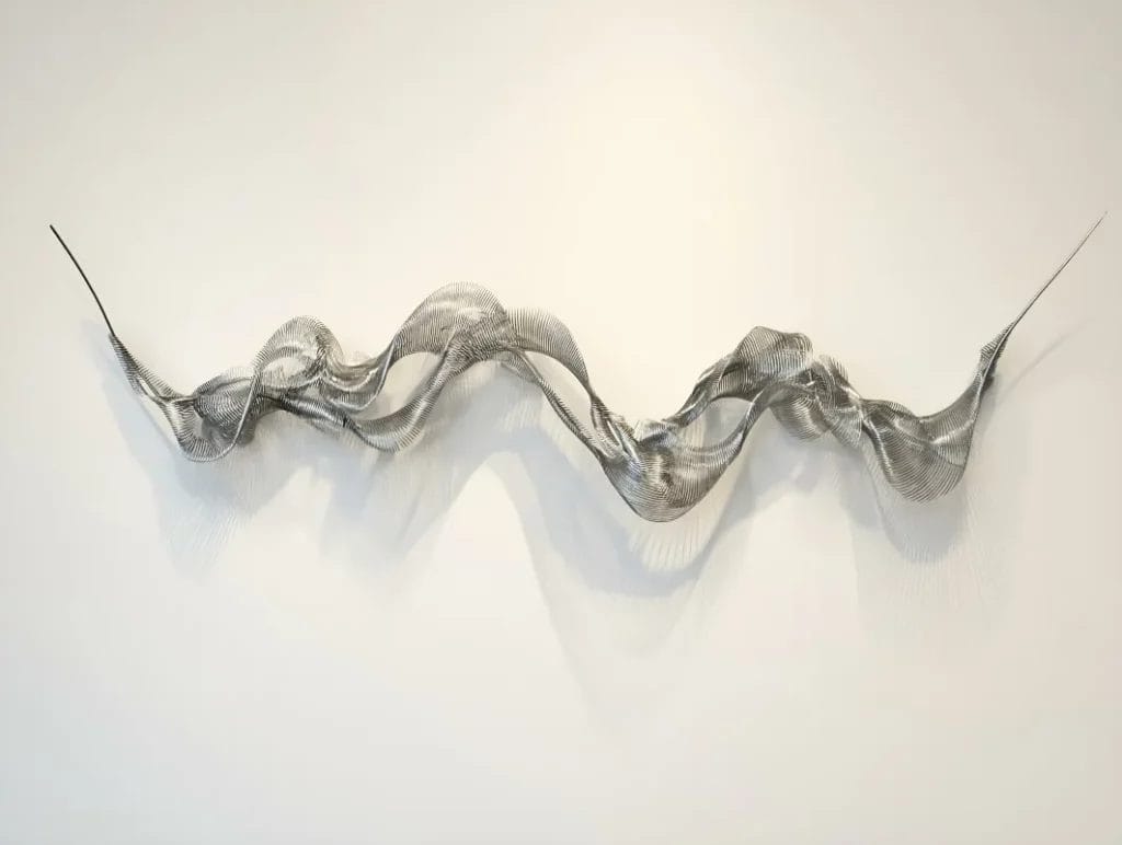 An abstract metal sculpture consisting of waves, twists, and two pointed ends is displayed against a white wall.