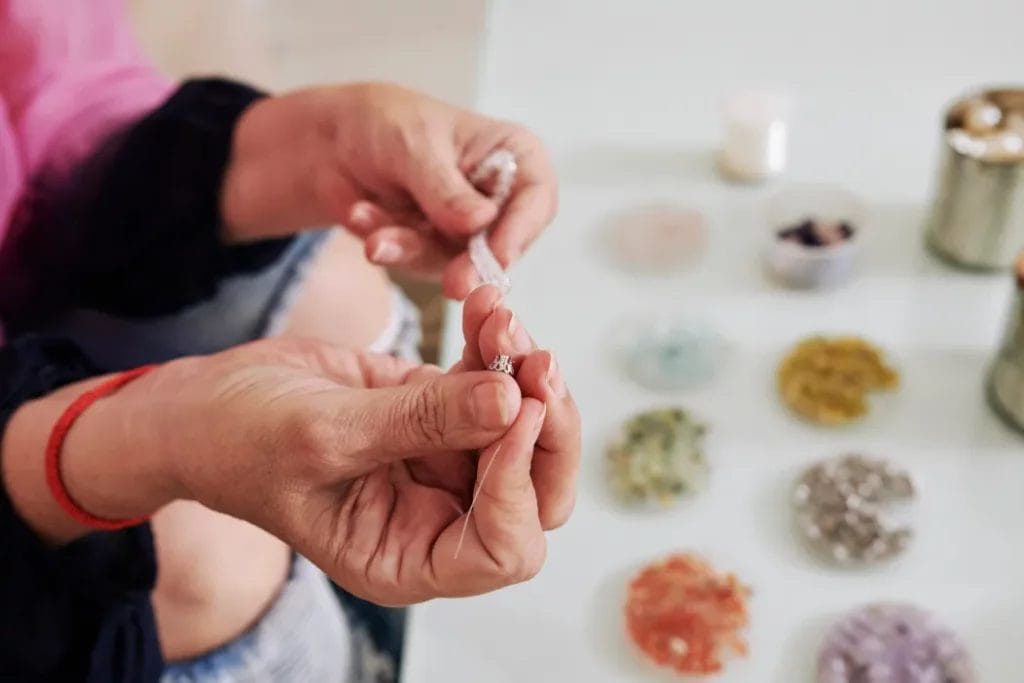 A close-up of a jewelry maker's hands as they thread stone beads on a wire strand above a table with trays of various beads in different colors.