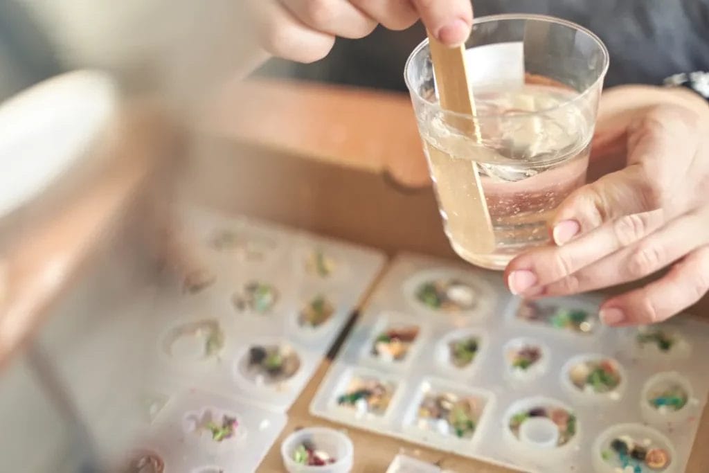 A jewelry maker stirs resin with a wooden stick in a clear cup above white trays of jewelry pieces.