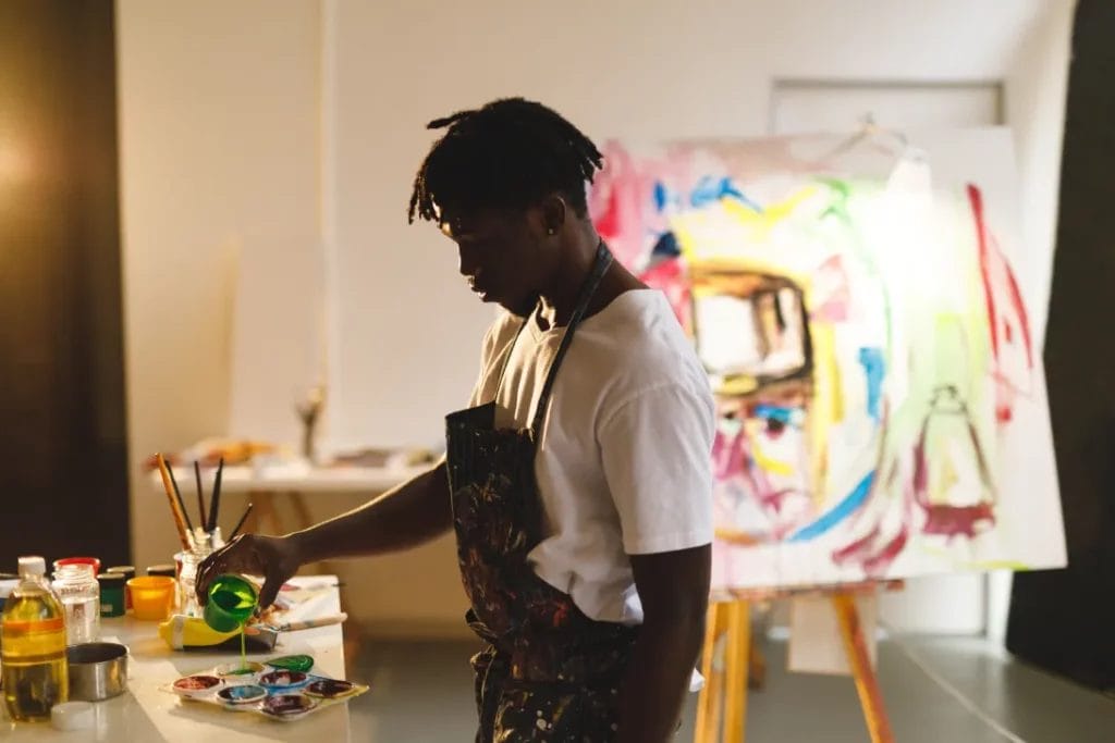 An artist wearing a white T-shirt and an apron pours paint into trays at a table in an art studio with a large colorful canvas in the background.