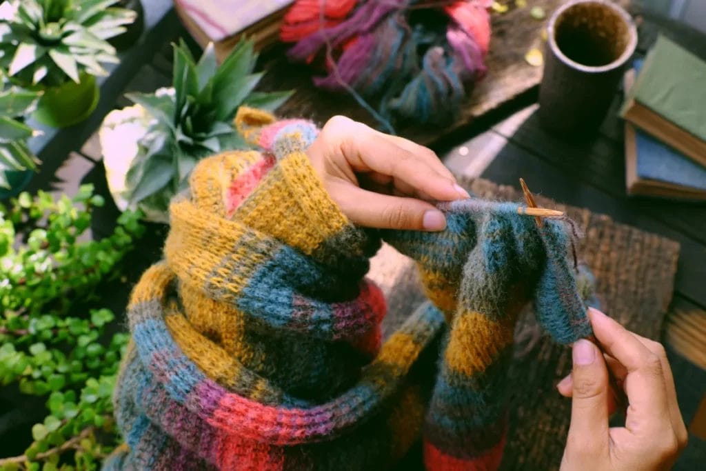 A close-up image of hands knitting a scarf with yellow, blue, red, and pink yarn above a table with various plants, books, and a mug.