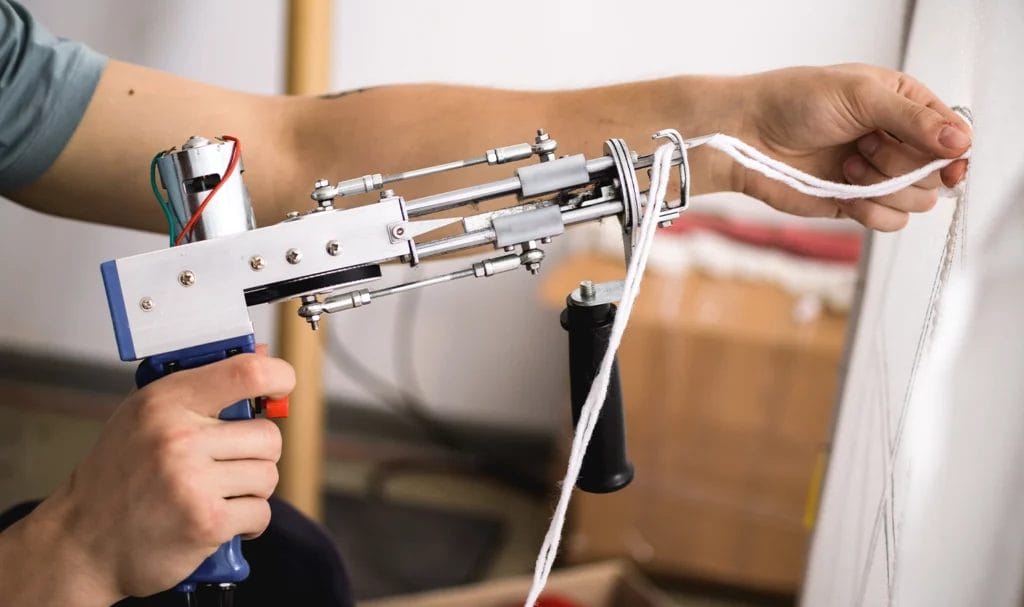 Close-up view of an artisan holding a thread and rug tufting machine near his canvas.