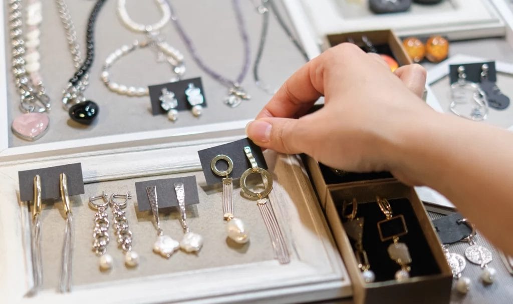 A close-up look of a woman's hand delicately selecting a pair of handmade earrings from a vendor's booth, showcasing an array of artisanal jewelry in the background.
