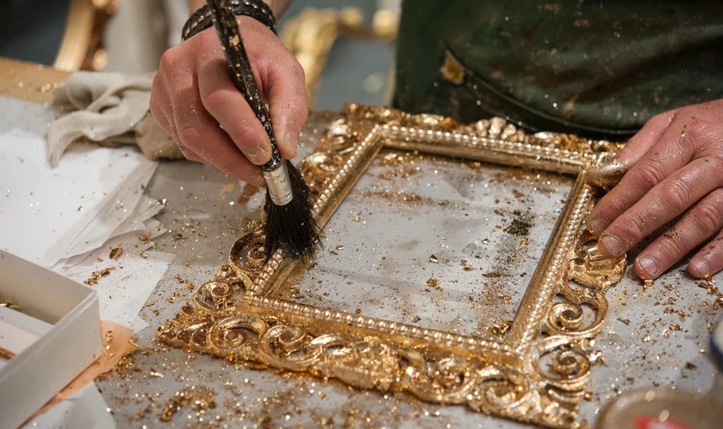 A master craftsman uses the gilding process technique to apply gold leaf to a handmade wood picture frame.