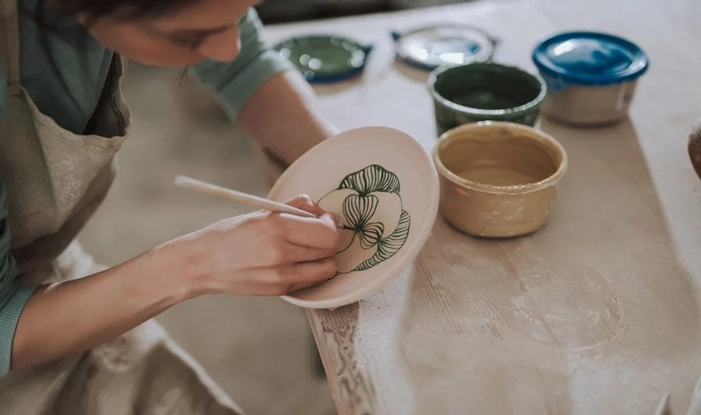 Young artist in apron painting a ceramic plate on a wooden workshop table.