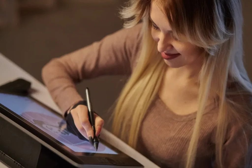 An artist with long blonde hair wearing a brown top and a black glove draws using a pen stylus on an artist's tablet.