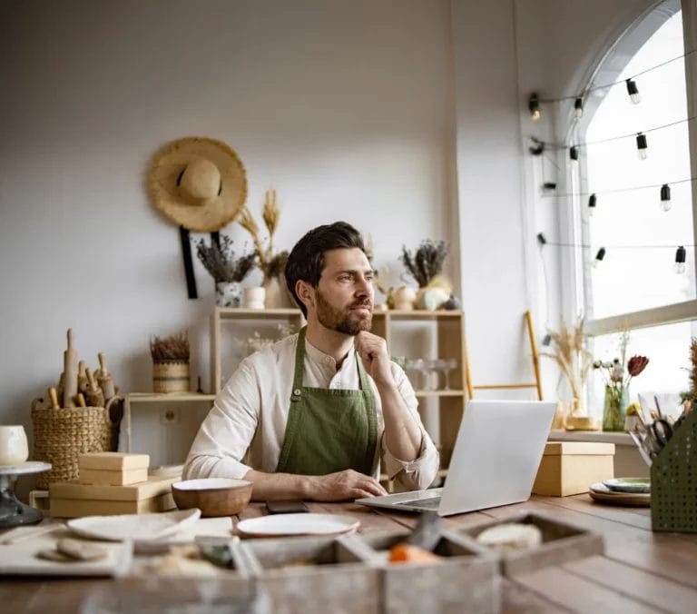 An artist wearing a white long-sleeve shirt and a green apron holds his chin while working at a table with a laptop, shipping boxes, ceramics, and tools in an art studio.