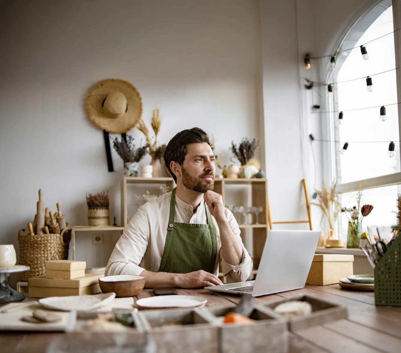 An artist wearing a white long-sleeve shirt and a green apron holds his chin while working at a table with a laptop, shipping boxes, ceramics, and tools in an art studio.