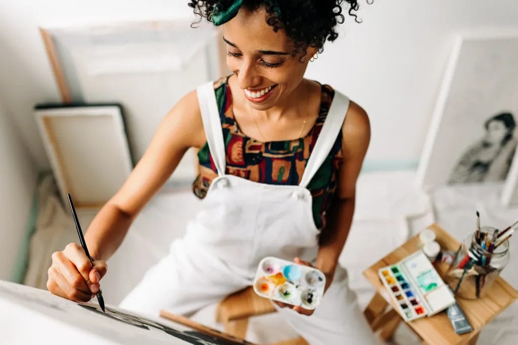 An artist wearing an abstract tank and white overalls paints a canvas and holds a palette with watercolor paints in a studio.