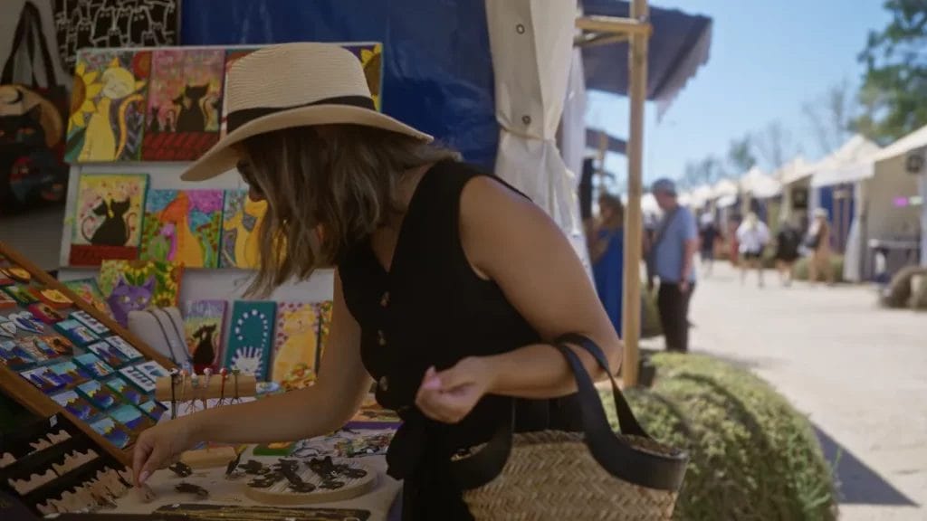 A woman wearing a straw hat and a black vest shops at an art vendor booth at an outdoor market.