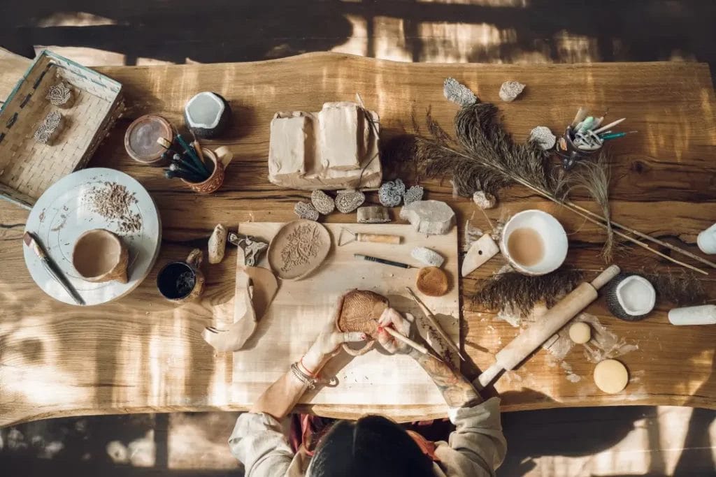 An artist sits at a wooden table covered with various art tools and uses a sharp tool to create a pattern on a ceramic piece.