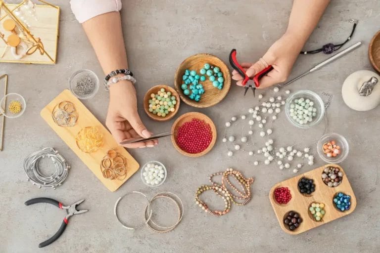 A top-view image showing a jewelry maker's hands holding red pliers and silver tweezers above various trays of beads, wires, and other jewelry materials on a gray table.