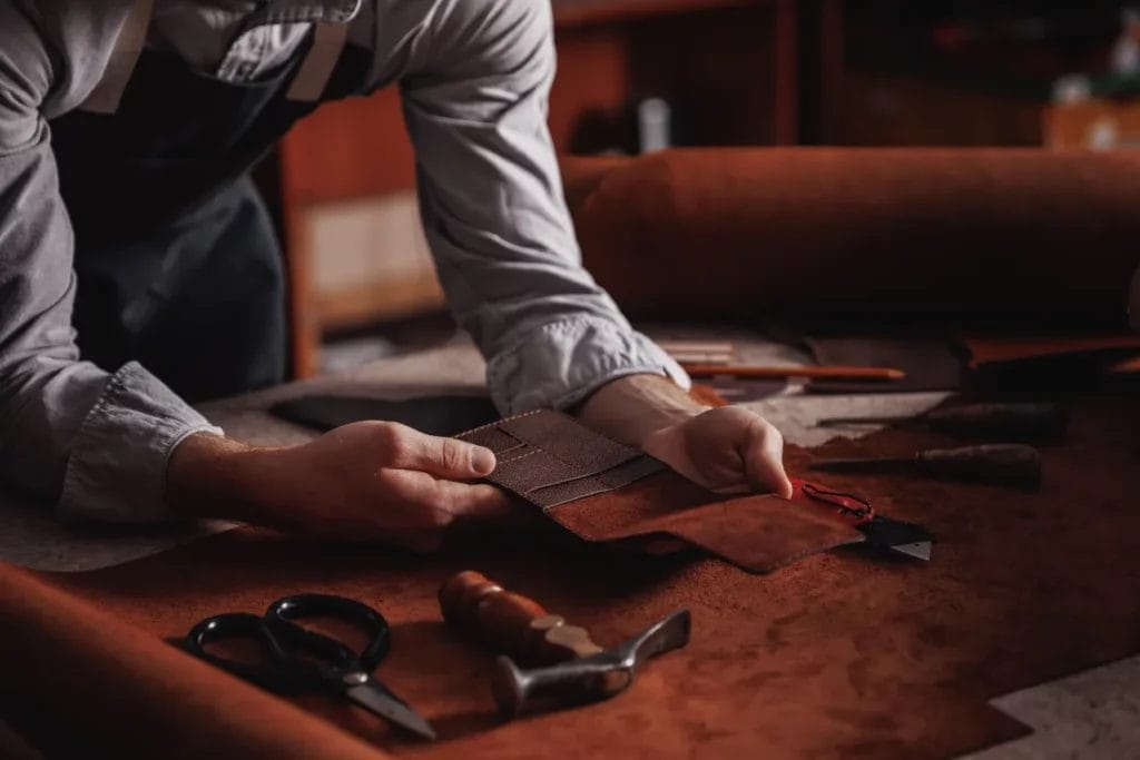 A leatherworker wearing a gray shirt and a navy apron examines a leather wallet over a brown worktable with scissors, a hammer, and other tools.